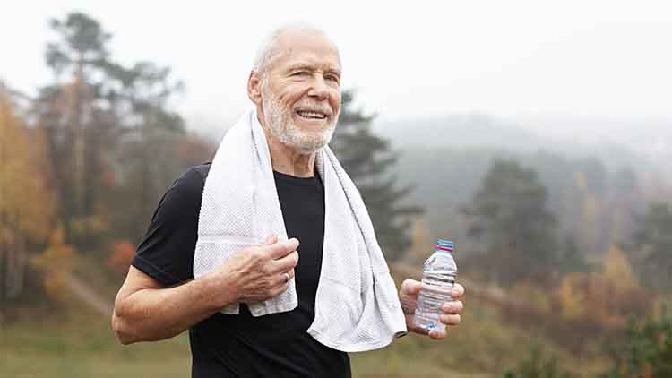 Exhausted elderly male with gray hair and beard drinking water after outdoor exercise and wiping sweat with towel around his neck