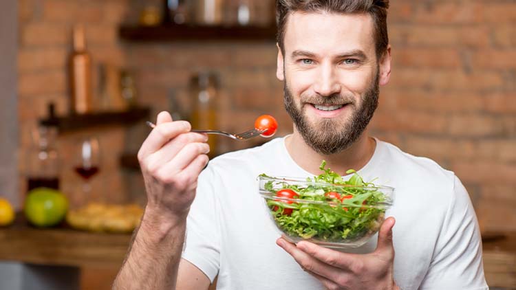 Handsome bearded man in white t-shirt eating salad with tomatoes in the kitchen. Healthy and vegan food concept