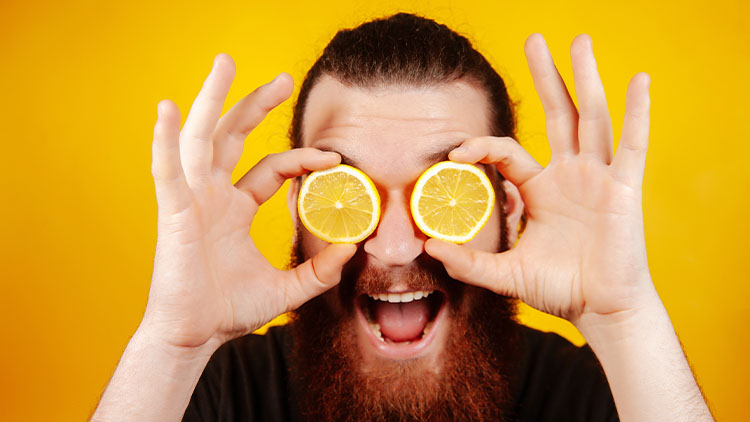 Happy bearded hipster man with sliced lemon on eyes over orange background. Man with long beard hold lemon. Fruit and healthy organic food. Citrus vitamin.