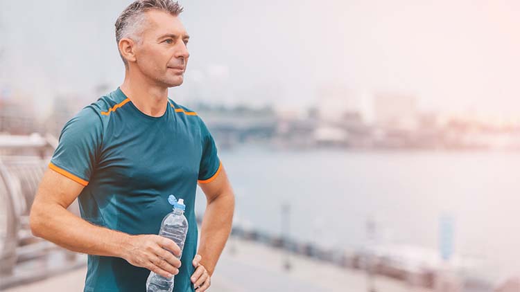 Portrait of healthy athletic middle aged man with fit body holding bottle of refreshing water, resting after workout or running. on the riverside.