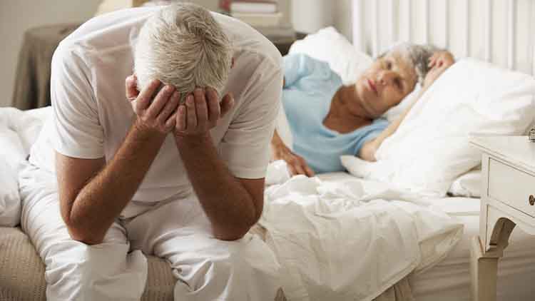 Worried Senior Man Sits On Bed Whilst Wife Sleeps