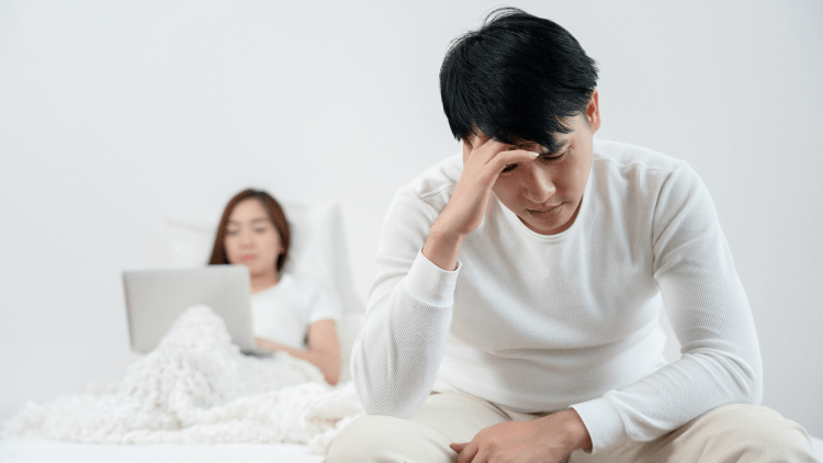 Man sat at edge of bed stressed while partner is in bed using laptop