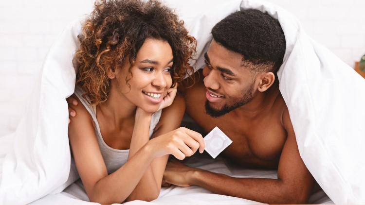 Young couple smiling under sheets holding condom